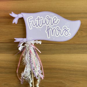 May include: A light purple banner with the words "Future Mrs." in white script, accented with diamond illustrations. The banner is adorned with a decorative tassel of white, pink, and lavender ribbons and a wooden stick.