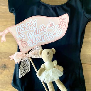 May include: A pink and white banner with the text "first day of dance!" and pink ballet shoe illustrations. A small teddy bear wearing a white tutu is in the foreground.