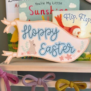 May include: A pink and blue banner with the text "Hoppy Easter" and a small brown bunny rabbit illustration. The banner is decorated with pink and blue flowers and green leaves.