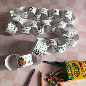 May include: A white paper chain with the text "Days to Halloween" and Halloween-themed illustrations. A box of Crayola crayons and several crayons are scattered around the chain. The chain is on a pink and white checkered surface.