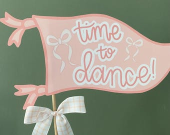 Op de afbeelding: Een roze banner met de tekst "time to dance!" in witte letters. De banner heeft aan beide uiteinden een roze strik en is bevestigd aan een houten stok met een roze-witte ruitjesstrik.