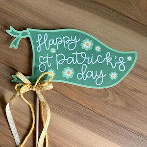 以下が含まれることがあります： 白い筆記体で「Happy St. Patrick's Day」と書かれた緑色のバナー。バナーは白と黄色の花で飾られており、黄色の水玉模様のリボンが付いた木の棒に付けられています。
