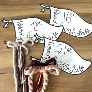 Könnte beinhalten: Drei weiße Wimpelflaggen mit schwarzer Beschriftung. Die Flaggen zeigen "1. Geburtstag", "2. Geburtstag" und "16. Geburtstag". Die Flaggen sind an Holzstäben mit buntem Garn befestigt.