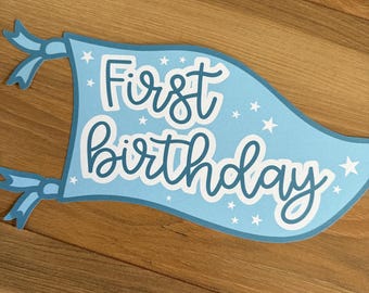 May include: A blue banner with white stars and the text "First birthday" in white outlined lettering.