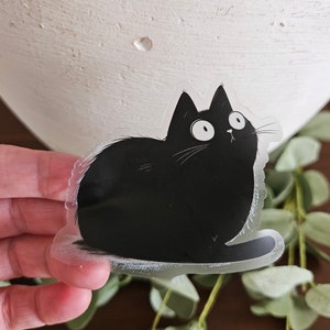 May include: A clear sticker featuring a cartoon black cat with wide eyes and a surprised expression. The cat is sitting, with its tail curled. The sticker is held in front of a white vase and green foliage.
