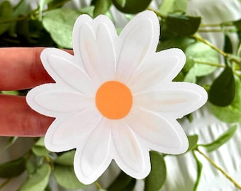May include: A white daisy sticker with an orange center. The sticker is on a white surface with green leaves.