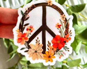 May include: A white sticker featuring a brown peace symbol encircled by a floral wreath. The wreath includes orange, red, and yellow flowers with green leaves. The sticker is held against a backdrop of green foliage.