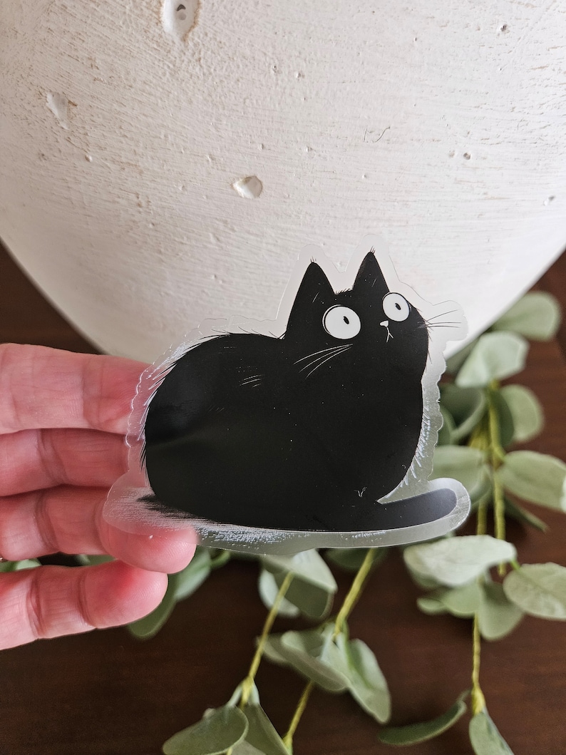 May include: A clear sticker featuring a cartoon black cat with wide eyes and a surprised expression. The cat is sitting, with its tail curled. The sticker is held in front of a white vase and green foliage.