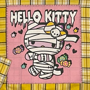 May include: A pink fabric patch with a white outline and the text "HELLO KITTY" in black. The patch features a cartoon image of Hello Kitty dressed as a mummy with a candy cane bow. The background is filled with hearts, stars, and candy.