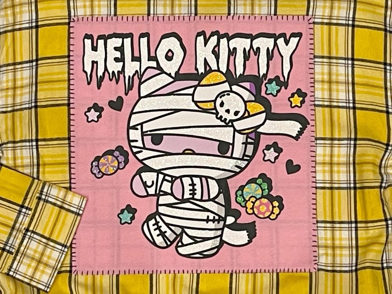 May include: A pink fabric patch with a white outline and the text "HELLO KITTY" in black. The patch features a cartoon image of Hello Kitty dressed as a mummy with a candy cane bow. The background is filled with hearts, stars, and candy.