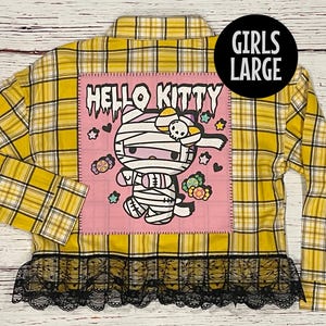 May include: Yellow and black plaid flannel shirt with a pink patch featuring a Hello Kitty mummy design. The shirt has a black lace trim.