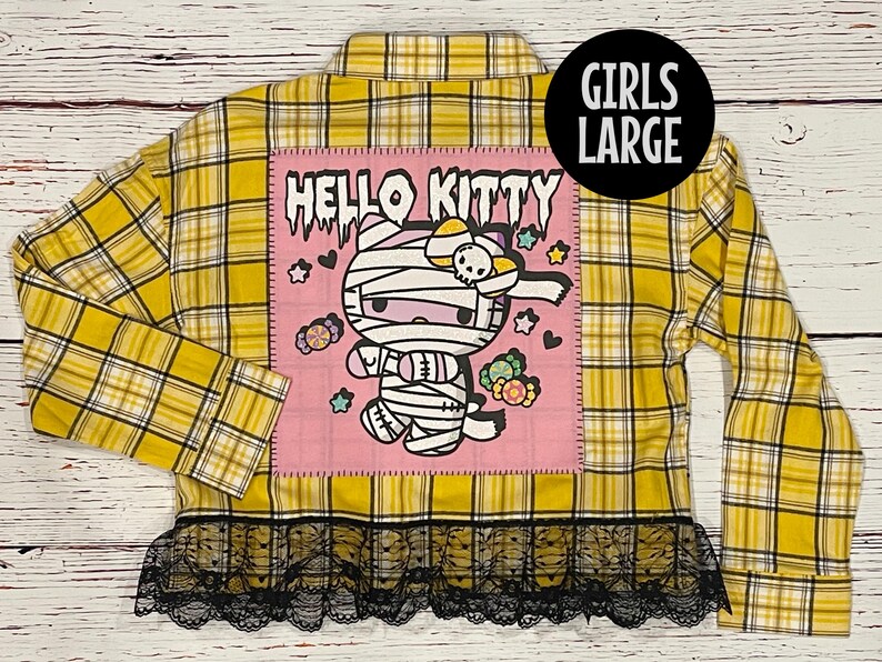 May include: Yellow and black plaid flannel shirt with a pink patch featuring a Hello Kitty mummy design. The shirt has a black lace trim.