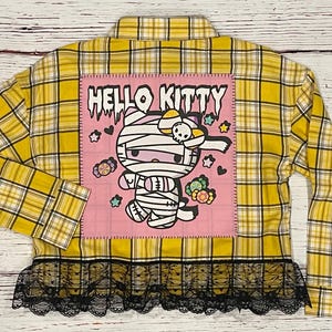 May include: Yellow and black plaid flannel shirt with a pink patch featuring Hello Kitty dressed as a mummy. The patch has the text "Hello Kitty" in black. The shirt has a black lace trim.