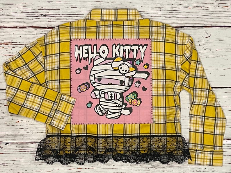 May include: Yellow and black plaid flannel shirt with a pink patch featuring Hello Kitty dressed as a mummy. The patch has the text "Hello Kitty" in black. The shirt has a black lace trim.