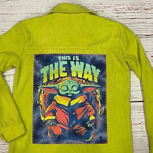 Op de afbeelding: Een limoengroen corduroy jack met een patch op de rug met daarop een cartoonfiguur met groene huid en grote oren. De patch zegt "This is the way" in een kleurrijk lettertype.