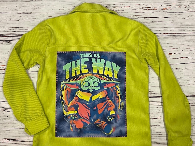 Op de afbeelding: Een limoengroen corduroy jack met een patch op de rug met daarop een cartoonfiguur met groene huid en grote oren. De patch zegt "This is the way" in een kleurrijk lettertype.