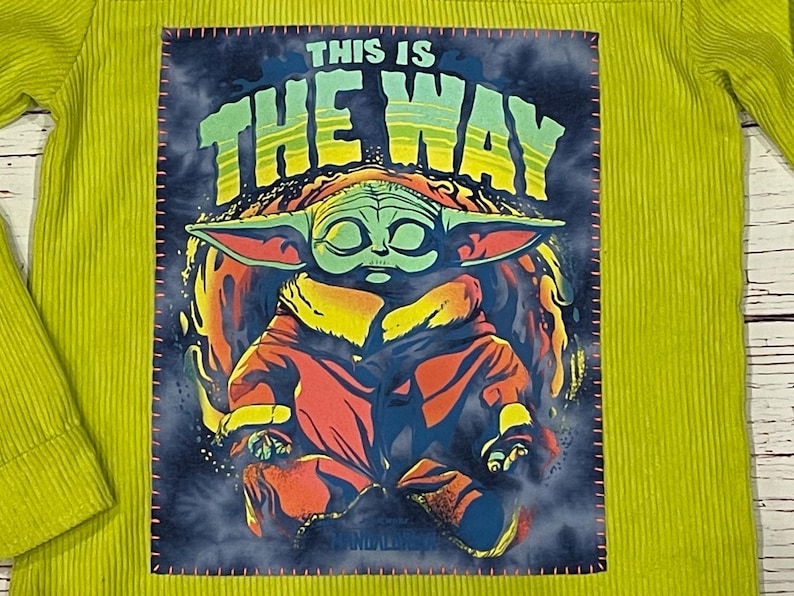 Op de afbeelding: Een geel corduroy shirt met een groene, gele en rode afbeelding van een personage uit de Star Wars-franchise, bekend als "Baby Yoda", met de tekst "This is the way".