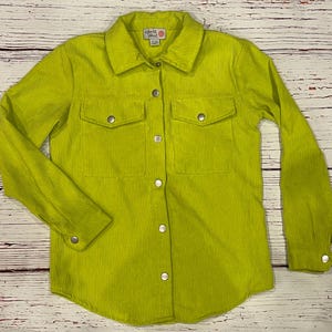 Op de afbeelding: Een limegroen corduroy overhemd met lange mouwen en twee borstzakken. Het shirt heeft een klein wit label met de tekst "Girls Like" erop.