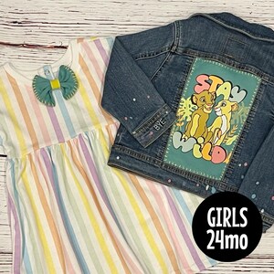 May include: A striped rainbow dress and a denim jacket with a patch that says "Stay Wild" and features a cartoon image of two lions. The jacket is for a 24-month-old child.