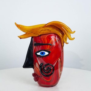 Pode incluir: Um vaso de vidro artístico vermelho e amarelo em forma de rosto. O vaso apresenta um rosto estilizado com um nariz preto, um olho azul e cabelo amarelo. Um objeto decorativo único.