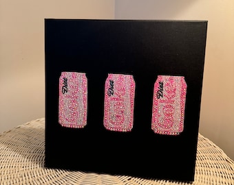 以下が含まれることがあります： 黒いキャンバスに、ピンク色の Diet Coke の缶が 3 つ描かれています。 各缶はキラキラ光るラインストーンで装飾され、「Diet」の文字がプリントされています。 缶は横に並んでいます。