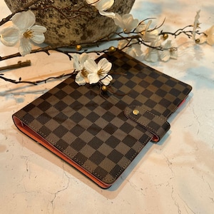 May include: A brown and black checkered pattern planner with an orange interior. The planner has a snap closure and is decorated with white flowers and branches.