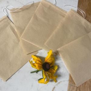 May include: Five beige tea bags with white string closures are arranged on a light-colored surface. A yellow and brown flower is placed near the tea bags. The tea bags are square-shaped and made of a translucent material.