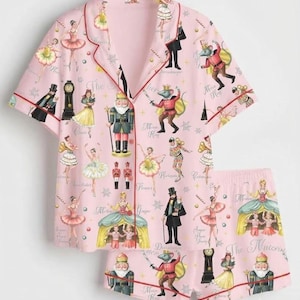 May include: A light pink short-sleeve button-down pajama shirt and matching shorts set featuring a repeating print of Nutcracker characters, including ballerinas, mice, and soldiers, with red piping along the collar and front closure.