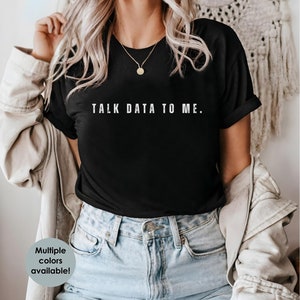 May include: A black t-shirt with the text "TALK DATA TO ME." printed in white on the front. The shirt is being worn by a person with long blonde hair and is paired with light blue jeans.