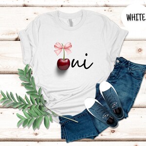 May include: White t-shirt with a red cherry and a pink bow on the front. The text "oui" is written in black cursive below the cherry. The shirt is laid out on a white wooden surface with a pair of blue jeans and white sneakers.