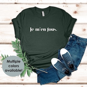 Puede incluir: Una camiseta verde oscuro con el texto "Je m'en fous." en letras blancas. La camiseta se muestra sobre un fondo de madera con un par de pantalones vaqueros azules y zapatillas blancas.
