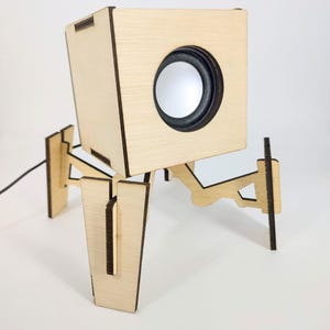 May include: A light-colored wooden speaker with a circular speaker and a black border. The speaker is mounted on a unique, geometric stand with four legs. A black power cord extends from the back of the speaker.