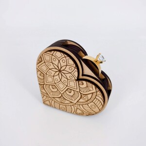 May include: A heart-shaped wooden ring box with an intricate mandala design. The box is light brown with darker brown edges. A gold ring with a clear gemstone sits in the open box, showcasing the ring's sparkle.