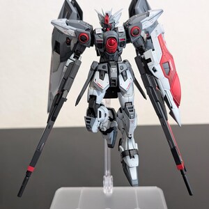 May include: A posable robot action figure featuring a color scheme of light gray, dark gray, and red. The figure has a central red chest orb, pointed head armor, and large wing-like attachments on the back. It is mounted on a clear plastic stand.