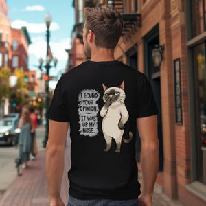 May include: A black t-shirt featuring a graphic of a Siamese cat picking its nose with the text I FOUND YOUR OPINION. IT WAS UP MY NOSE. The cat has a tan body, dark brown ears, face, and tail, and a grumpy expression on its face.