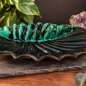 May include: A ceramic dish shaped like a shell with a glossy finish. The centre features a raised, textured ridge in vibrant emerald green and black, while the surrounding wide, scalloped edges are a deep, dark forest green with a smooth glaze.