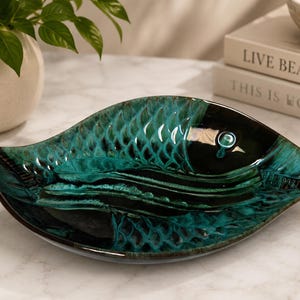Peut inclure: Un plat en céramique en forme de poisson avec une finition brillante dans des tons de bleu sarcelle profond, vert émeraude et noir. La surface présente un motif d'écailles texturé sur le corps et un détail d'œil circulaire près de la tête.