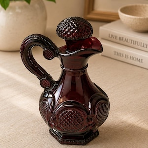 Pode incluir: Um decanter de vidro em tom bordô profundo com um padrão de diamante texturizado no corpo arredondado, uma tampa a condizer com topo facetado e uma pega curva. O vidro tem uma qualidade translúcida e acabamento polido com base hexagonal.