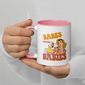 Puede incluir: Taza de cerámica blanca con borde y asa de color rosa. La taza presenta una ilustración de estilo retro de una mujer sosteniendo un megáfono con el texto "Support Babes" y "Babes" en una fuente naranja en negrita.