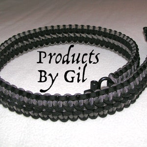 May include: Black and grey paracord strap with a black metal clasp. The strap is woven in a pattern and says "Products By Gil" in the center.