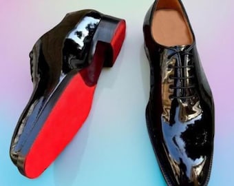 May include: Pair of glossy black dress shoes with red soles. The shoes have a classic lace-up design and a high-shine finish, reflecting the surrounding light. The soles are a vibrant red, contrasting with the black uppers.