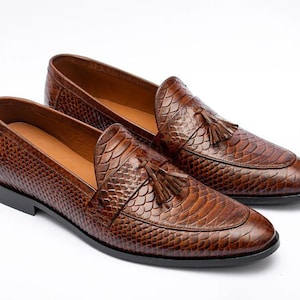 Peut inclure: Une paire de mocassins en cuir marron avec un motif peau de serpent et des accents de pompons.