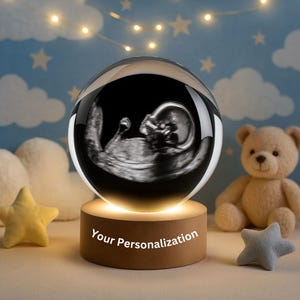 Può includere: Una sfera di cristallo trasparente con un'immagine ecografica di un feto, posta su una base di legno con la scritta "Your Personalization". Lo sfondo presenta un orsacchiotto, stelle e luci a catena.