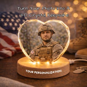 Puede incluir: Un recuerdo de cristal 3D en forma de corazón con una foto de un soldado, iluminado por luces cálidas. La base de madera tiene el texto "YOUR PERSONALIZATION". El texto "Turn Your Photo Into A 3-D Crystal Keepsake" está en la parte superior.