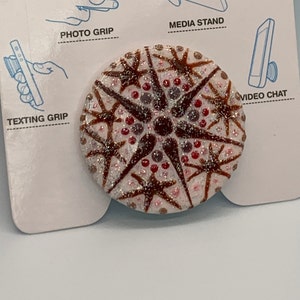 Puede incluir: Una agarradera de teléfono PopSocket blanca con un diseño de copo de nieve marrón y rojo. El PopSocket está en una tarjeta blanca con el texto "PopSockets Use Your Phone Better" e imágenes que muestran cómo usar el PopSocket para diferentes funciones del teléfono.