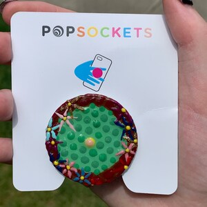 Puede incluir: Una agarradera de teléfono PopSockets verde y roja con un fondo blanco y un patrón de flores coloridas alrededor del borde. El logotipo de PopSockets es visible en el fondo blanco.
