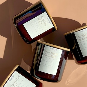 May include: Four brown glass candle jars with wooden lids. Each jar has a white label with gold lettering. The labels read "Santal Rose", "Vintage Chere", "Oakmoss & Amber", and "Cashmere Cream".