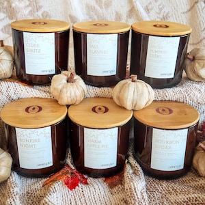 May include: Six dark brown glass candle jars with light bamboo lids, each featuring a white label with text including Cider Spritz, Fall Flannel, Spiced Pumpkin Creme, Bonfire Night, Warm Apple Pie, and Bourbon Sugar, surrounded by small dried pumpkins.