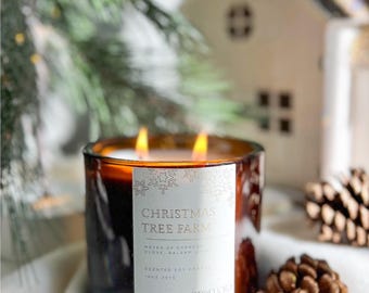 May include: A lit, scented soy candle in a brown glass jar. The label reads "Christmas Tree Farm" with snowflake accents. The candle has two wicks and is surrounded by pine cones and a miniature house, suggesting a holiday theme.