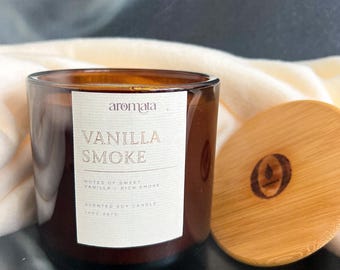 Puede incluir: Un tarro de vela de vidrio marrón con una etiqueta blanca que dice "aromata vanilla smoke" y "notes of sweet vanilla & rich smoke" y "scented soy candle 14oz 397g". La vela está sentada sobre una superficie oscura con una manta blanca y una tapa de madera con un logotipo.